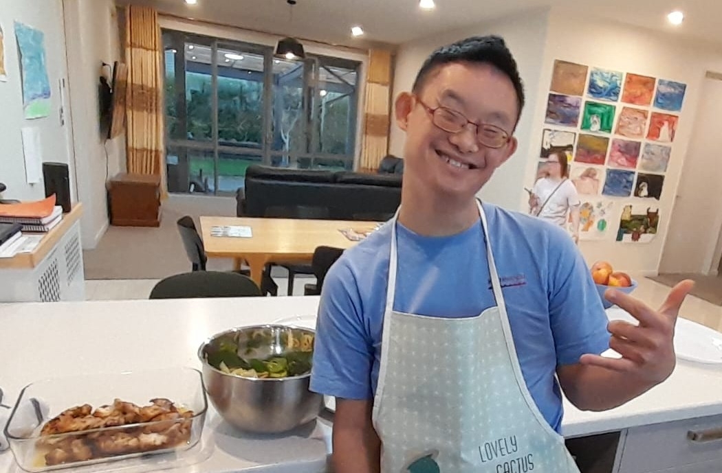 A happy young man grins as he stands in the kitchen beside food he's cooked.